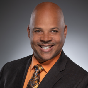 Professional headshot of Eric Smith, a smiling Black man in a dark suit, orange shirt, and patterned tie, against a gray background