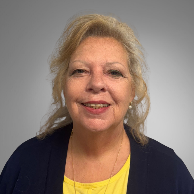 Professional headshot of Denise Wempe, a smiling middle-aged woman with shoulder-length blonde hair, wearing a navy blue cardigan over a yellow top, against a light gray background