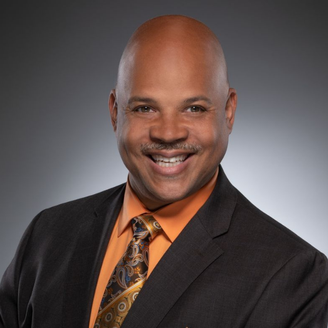 Professional headshot of Eric Smith, a smiling Black man in a dark suit, orange shirt, and patterned tie, against a gray background